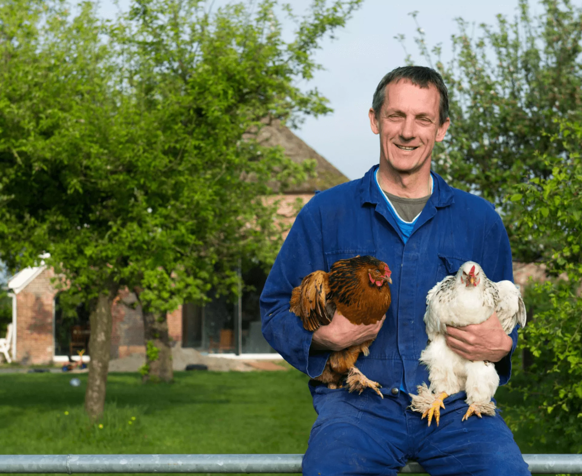 Man met twee kippen in zijn armen voor een boerderij.