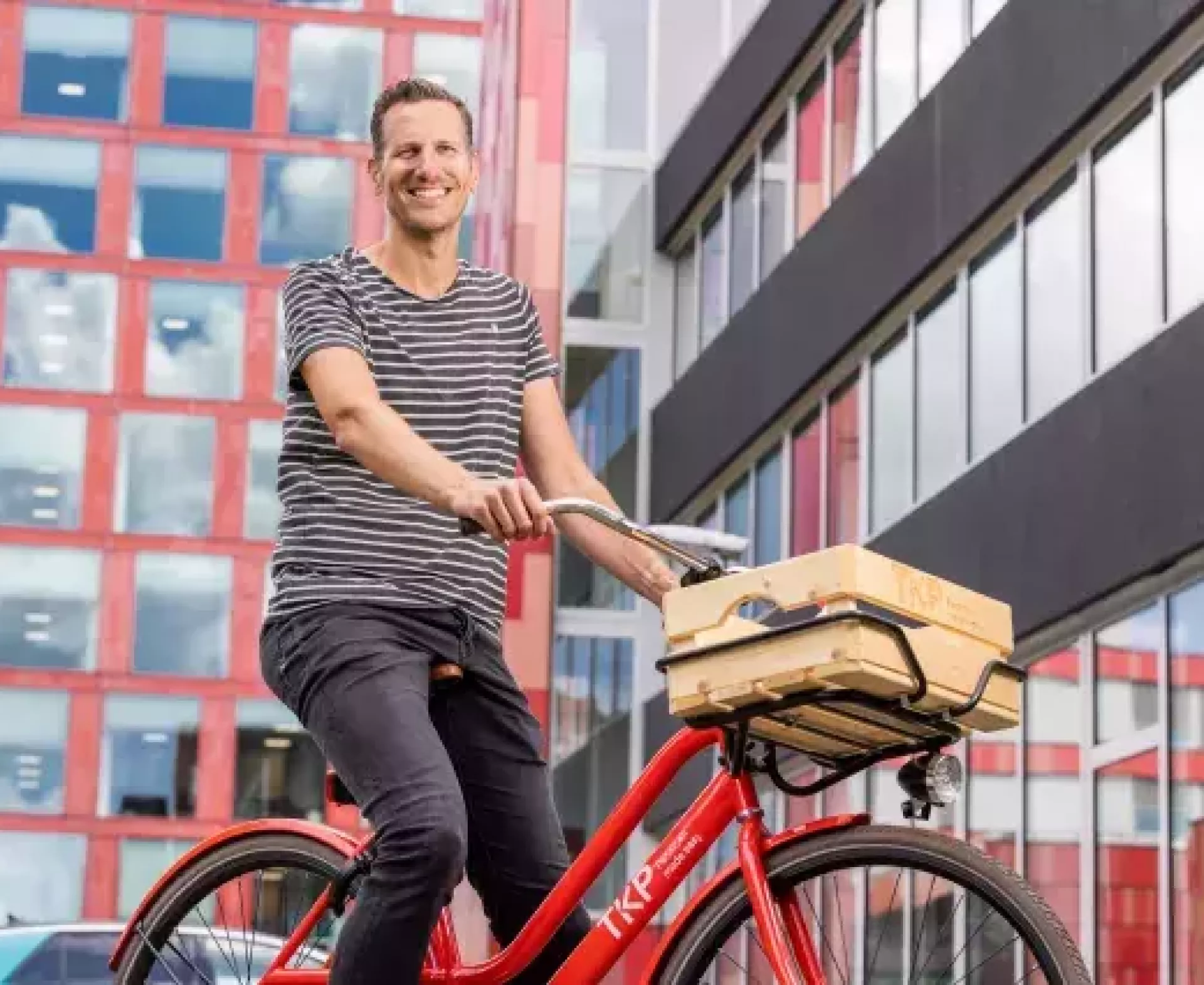 Een man op een rode fiets met een houten krat voor het gebouw van TKP.