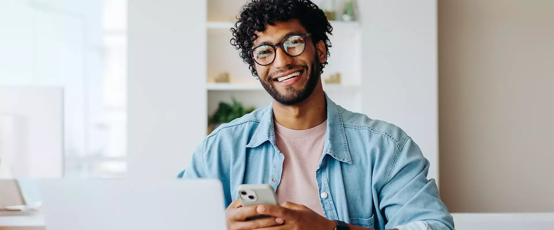 Een glimlachende man met bril zit achter een laptop en houdt een smartphone vast.