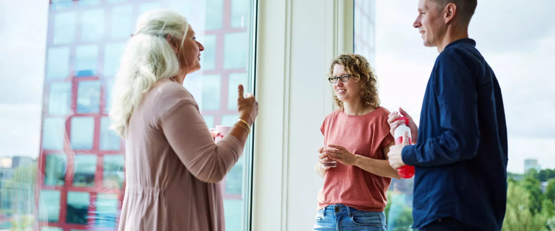 Drie mensen in gesprek bij een raam met uitzicht op een gebouw.