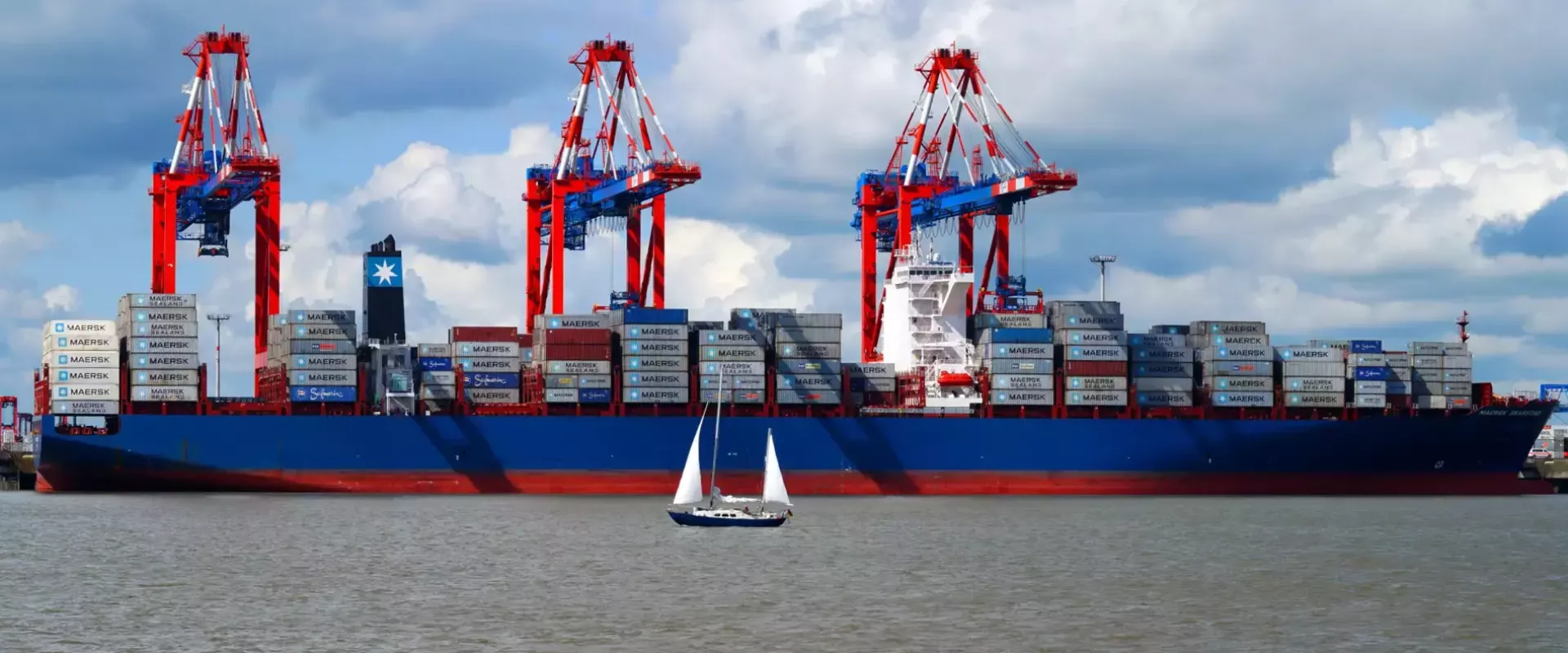 Een groot containerschip in de haven met een zeilboot op de voorgrond.