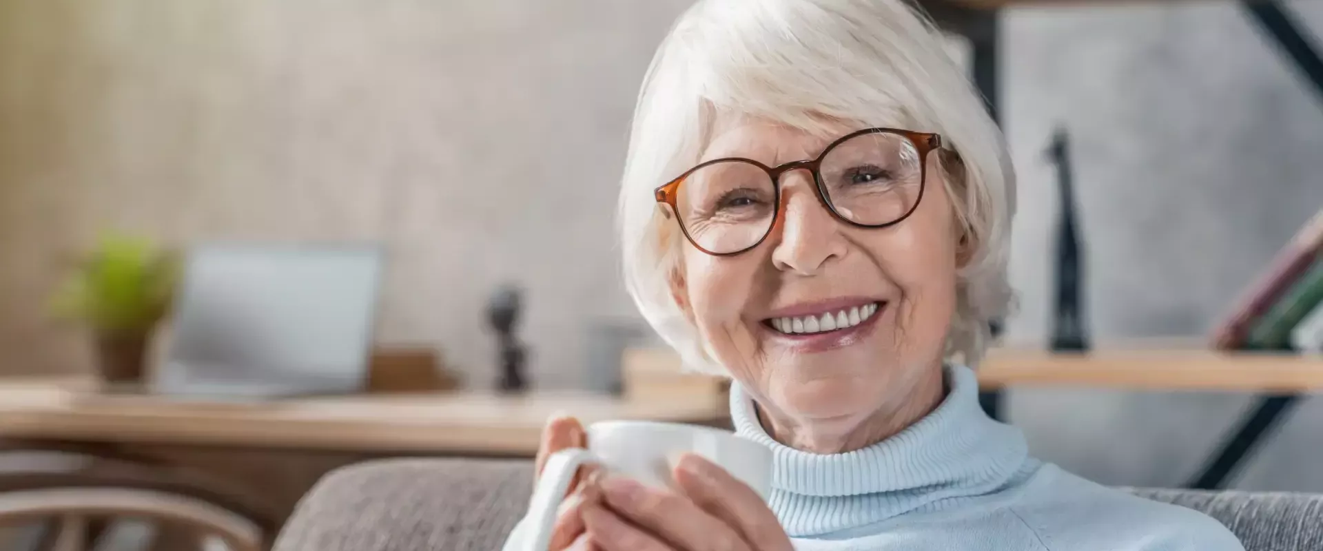 Een oudere vrouw glimlacht met een kopje koffie in de hand.