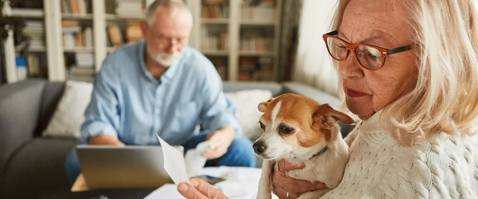Oudere vrouw met hondje leest een brief, terwijl een man op de achtergrond werkt.