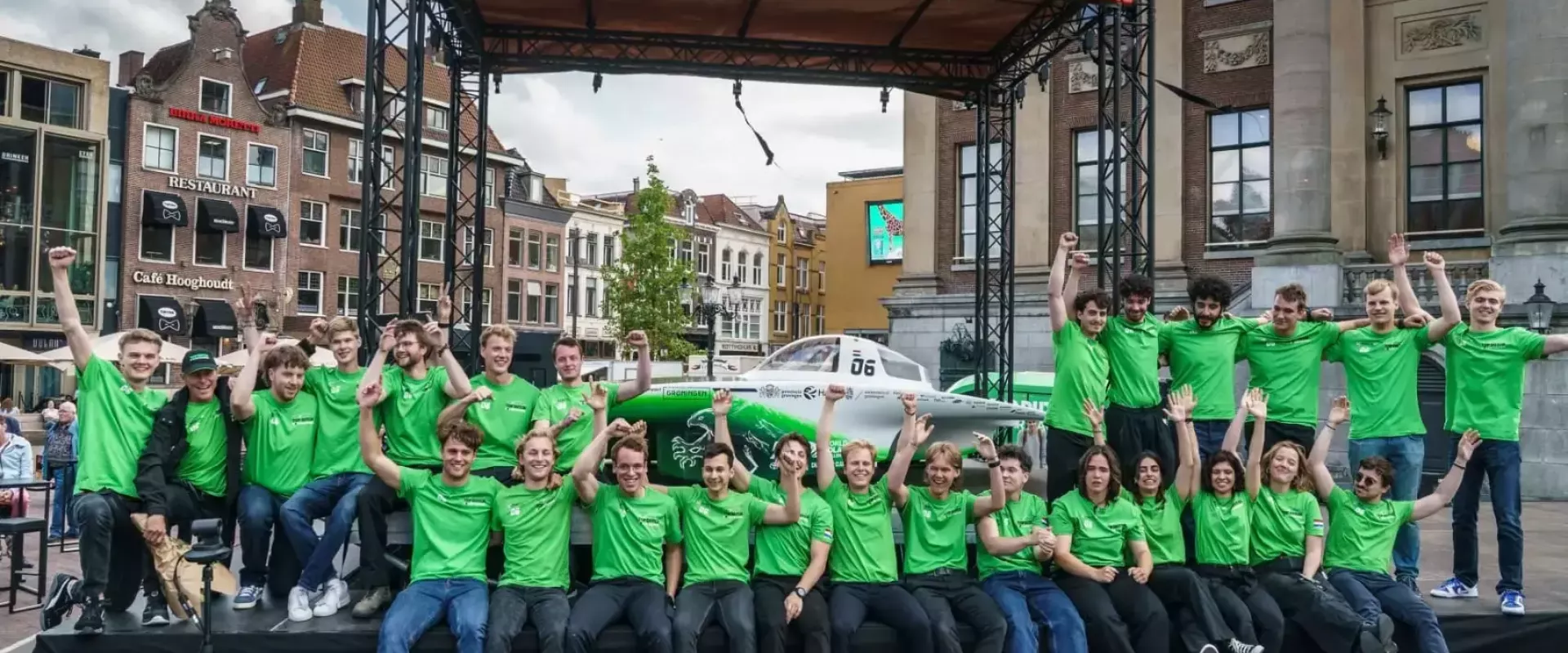 Groep mensen in groene shirts voor een zonneauto op een podium buiten.