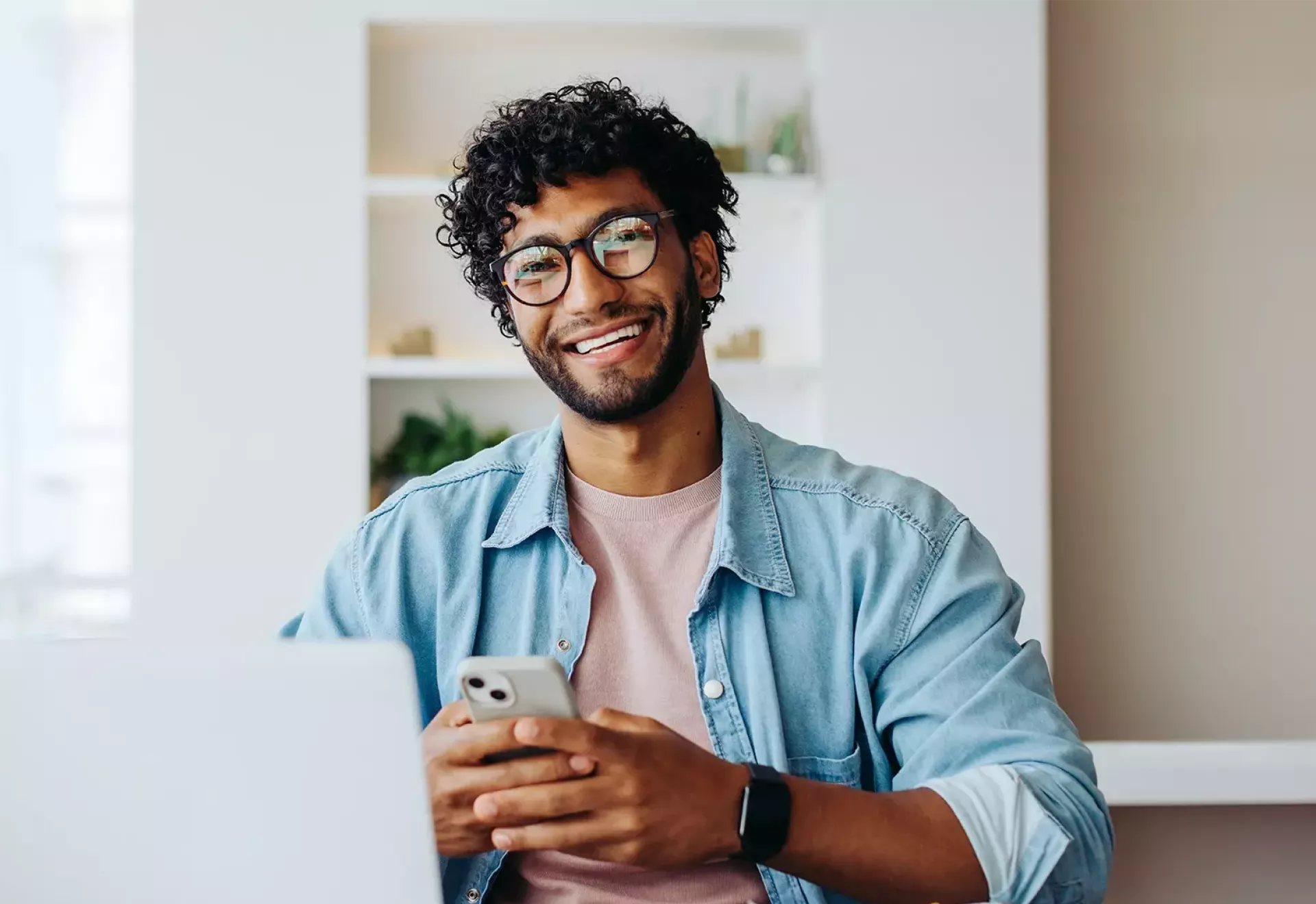 Een glimlachende man met bril zit achter een laptop en houdt een smartphone vast.