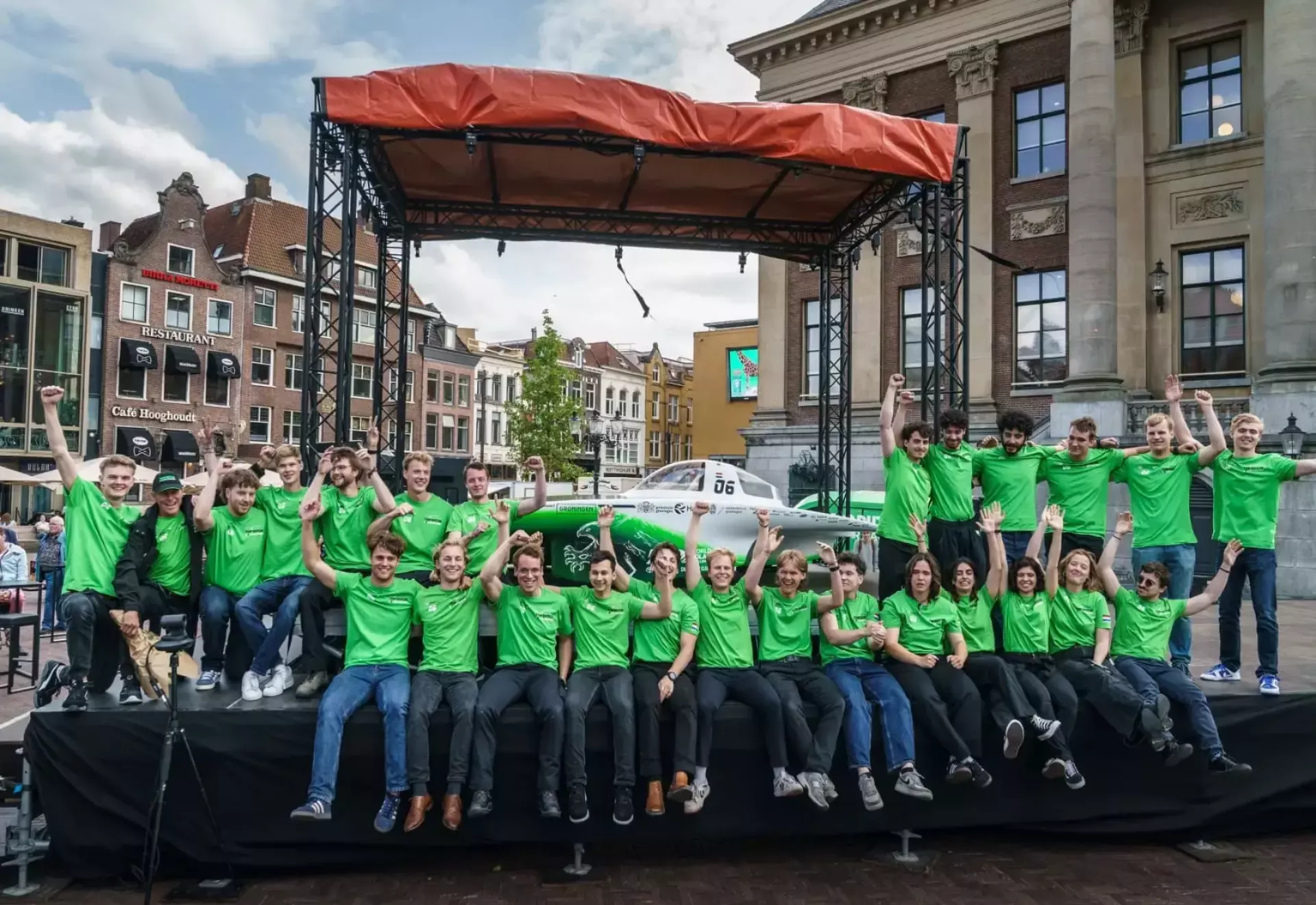 Groep mensen in groene shirts voor een zonneauto op een podium buiten.