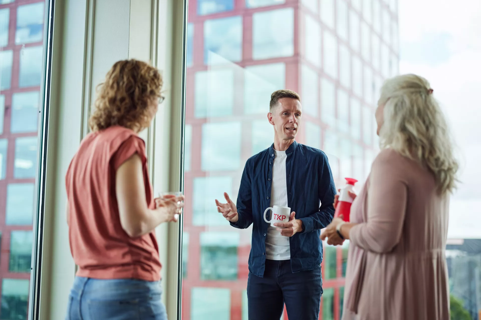 Drie mensen in een gesprek met koffiemokken voor een groot raam met uitzicht op een gebouw.