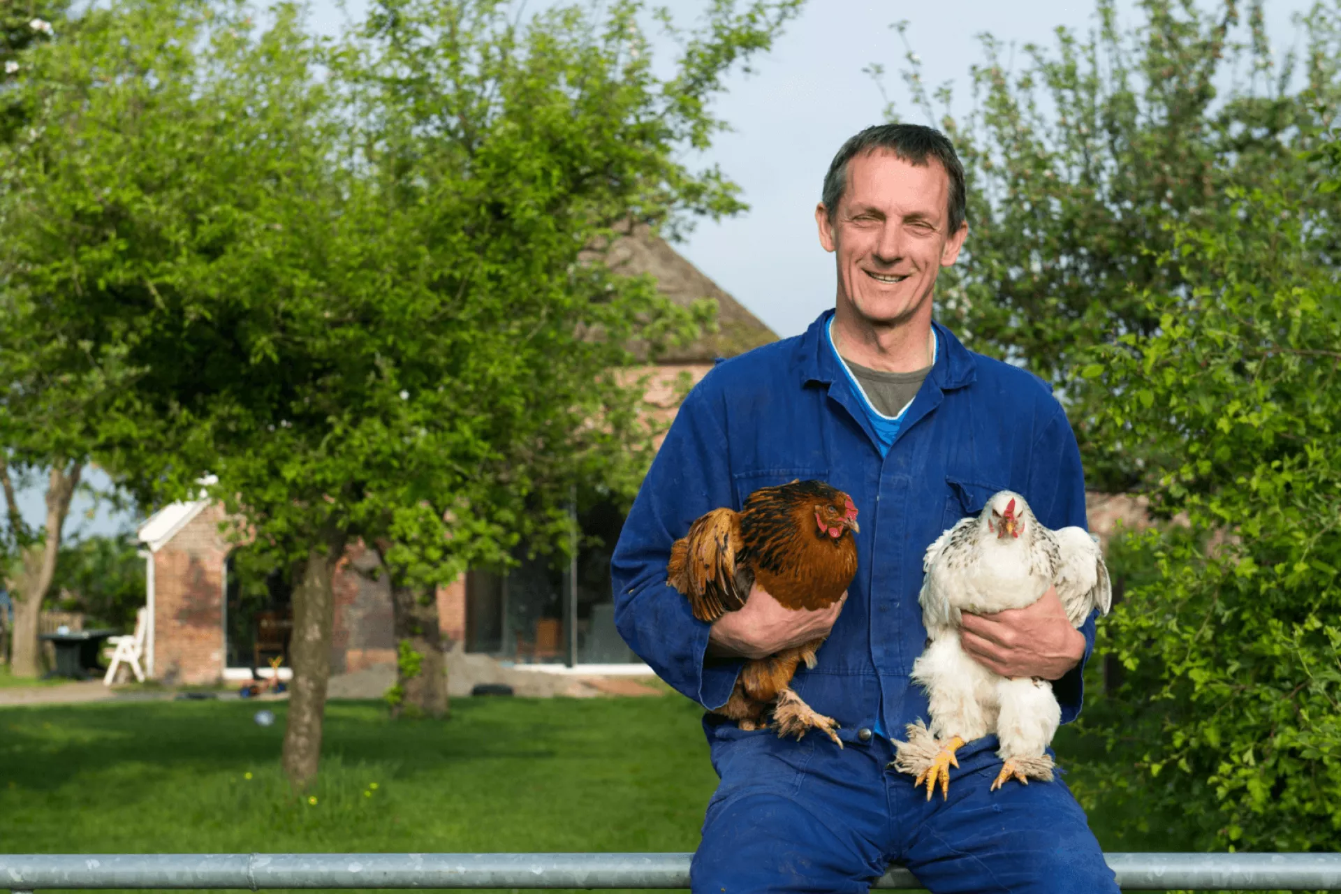 Man met twee kippen in zijn armen voor een boerderij.