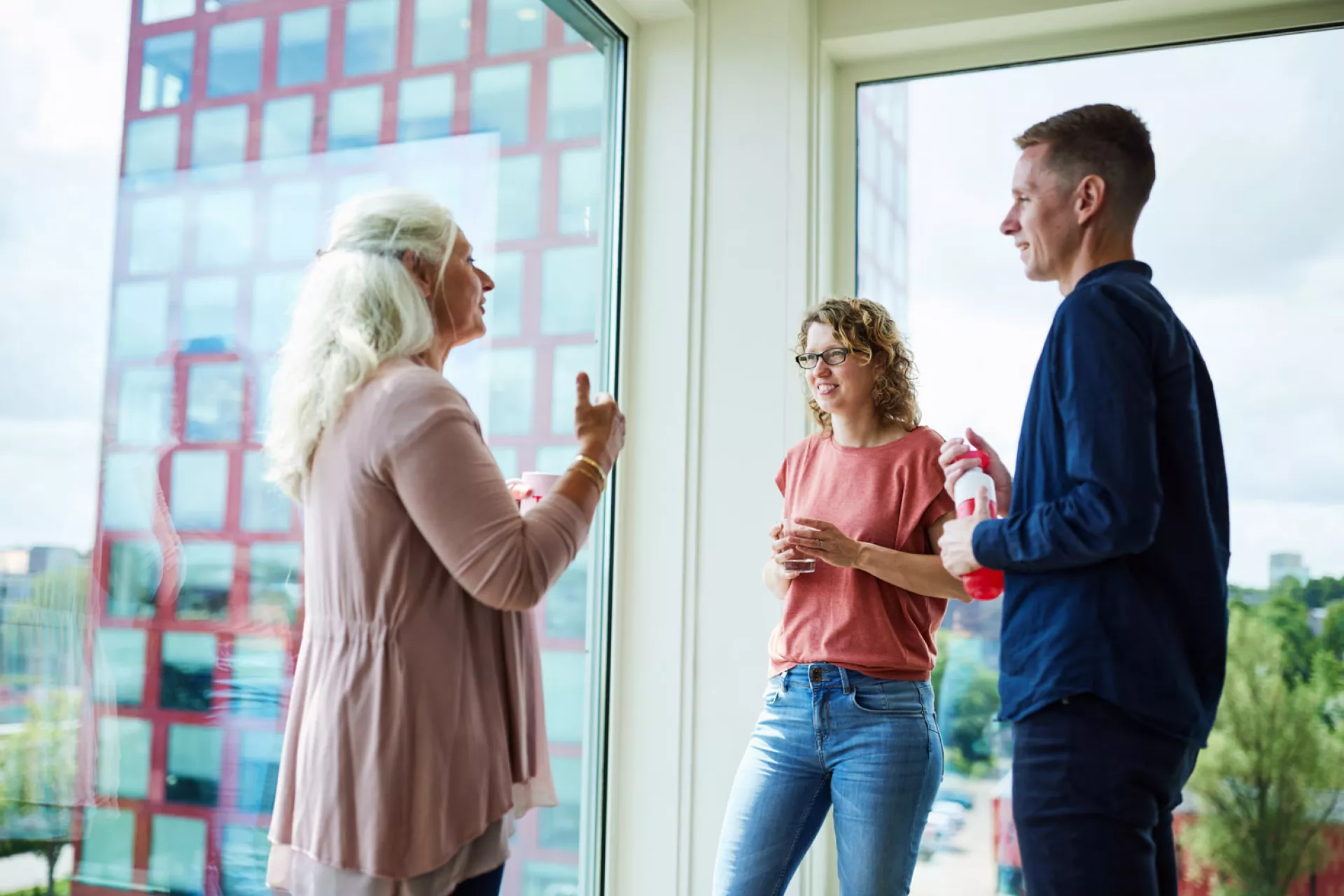 Drie mensen in gesprek bij een raam met uitzicht op een gebouw.