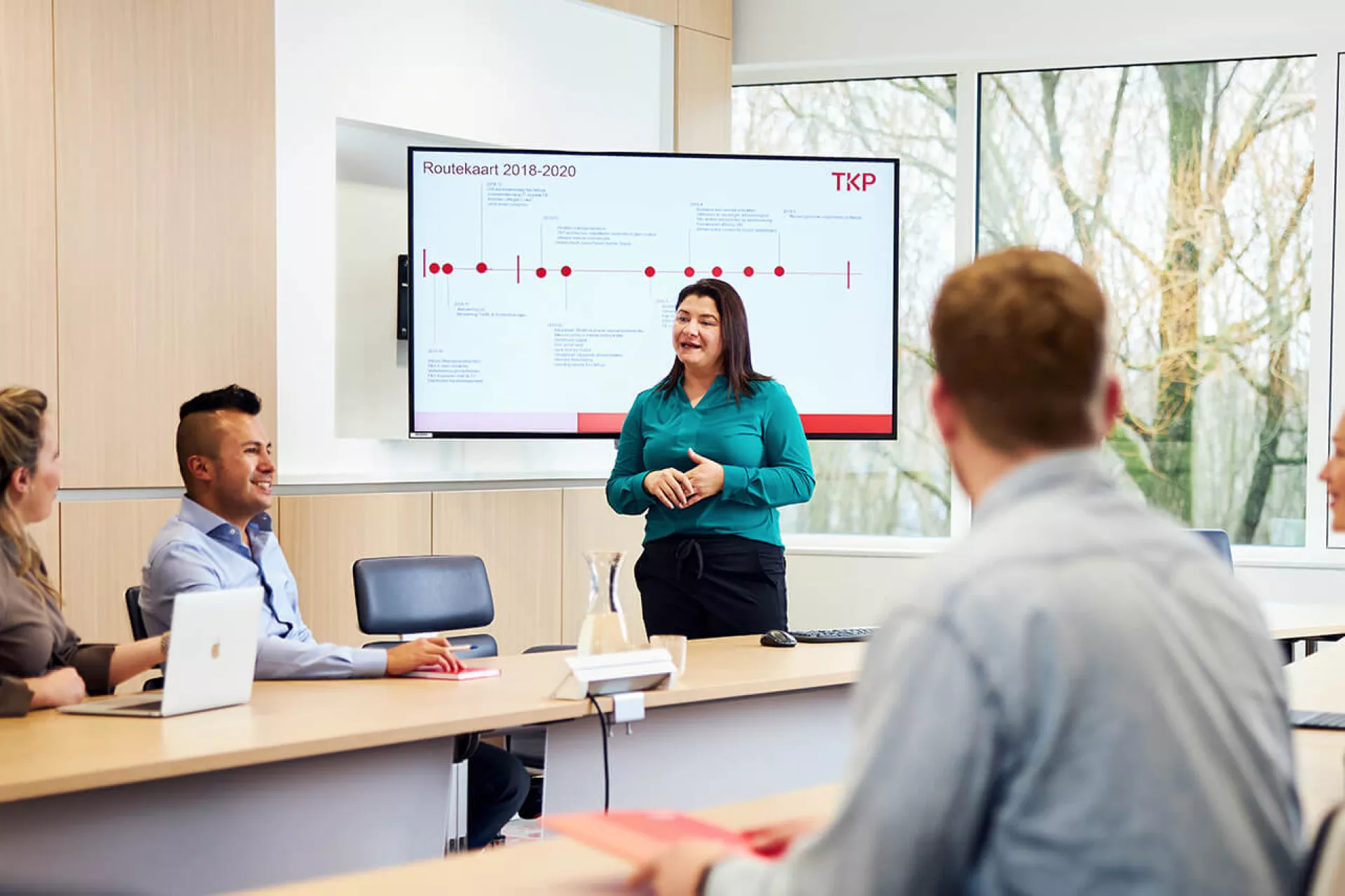Vergadering in een conferentieruimte met presentatrice voor een scherm.