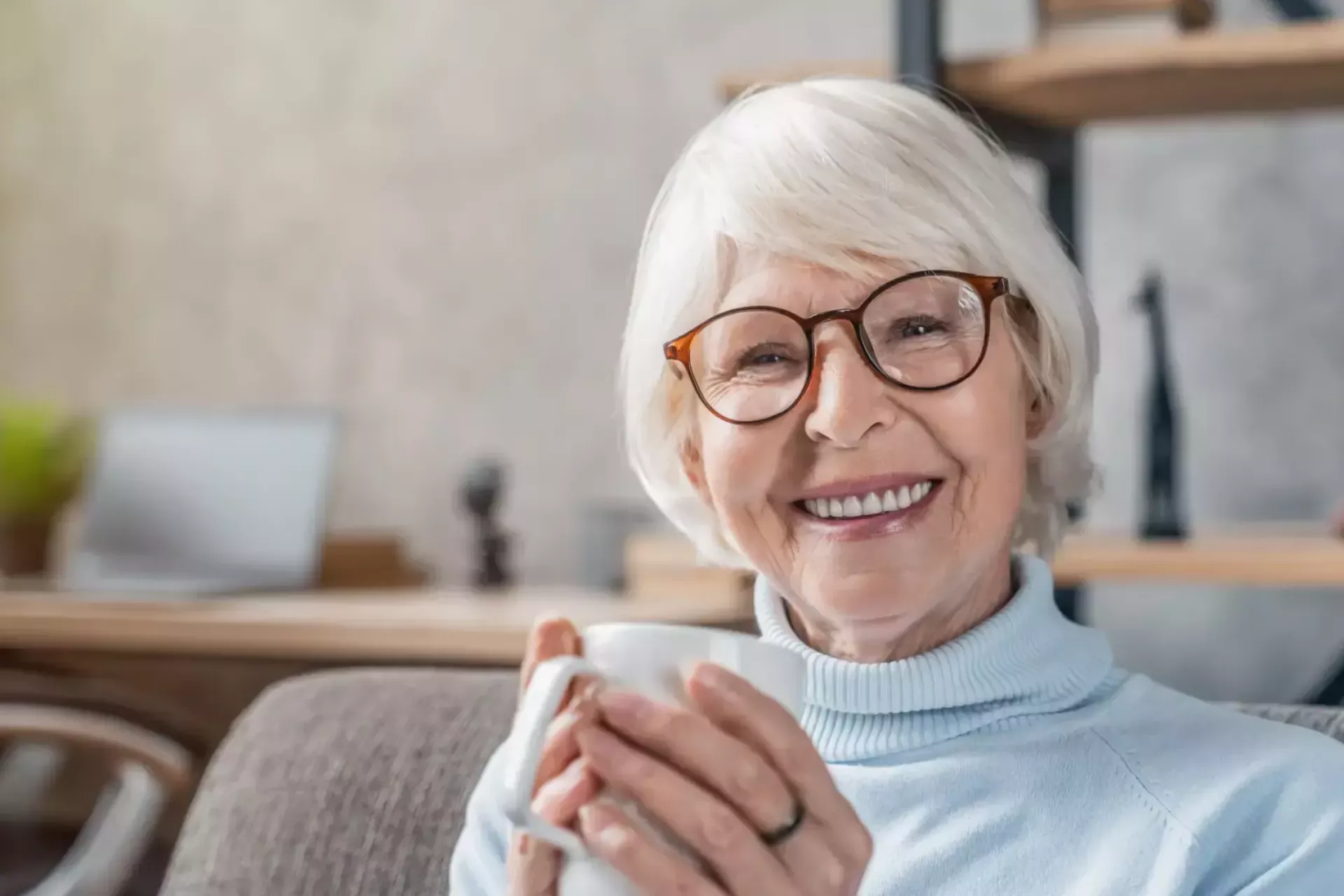 Een oudere vrouw glimlacht met een kopje koffie in de hand.