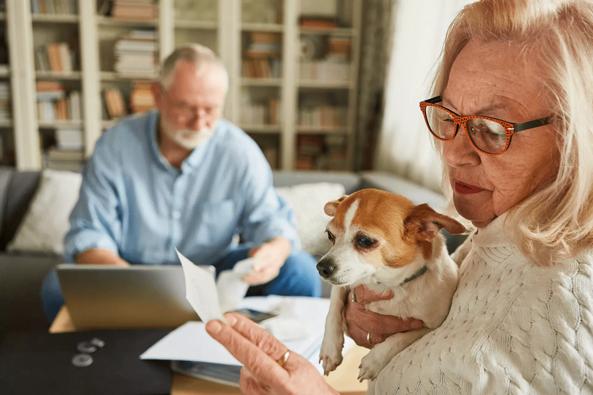 Oudere vrouw met hondje leest een brief, terwijl een man op de achtergrond werkt.