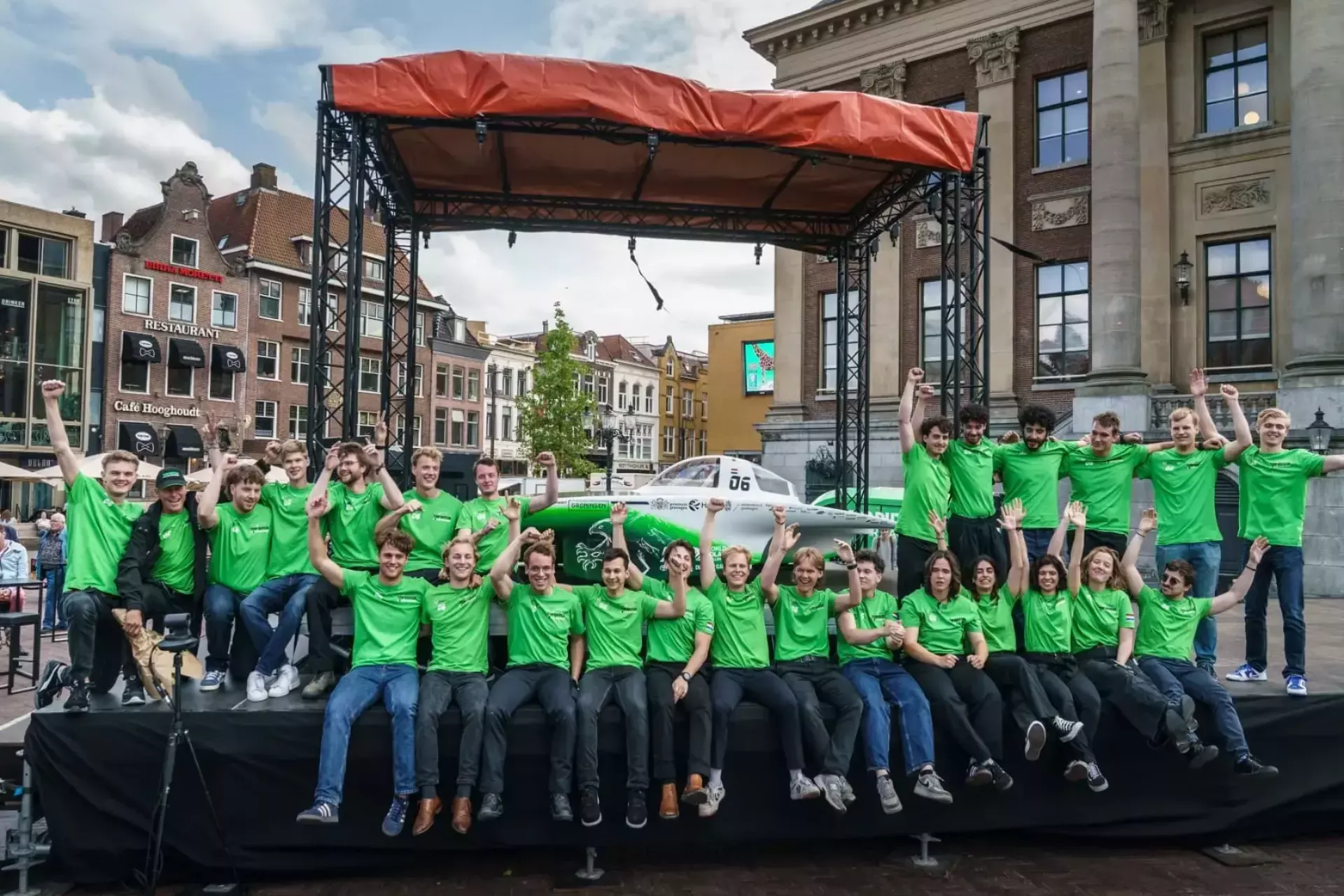Groep mensen in groene shirts voor een zonneauto op een podium buiten.