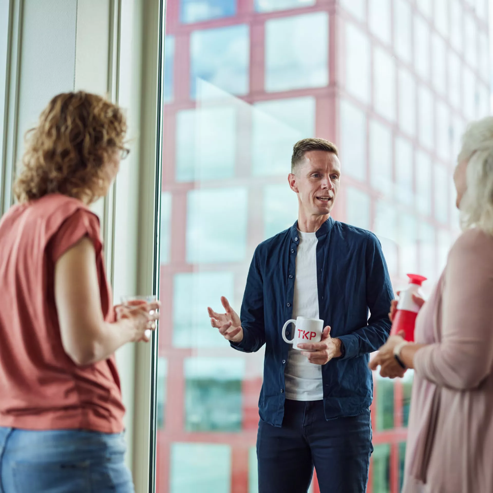 Drie mensen in een gesprek met koffiemokken voor een groot raam met uitzicht op een gebouw.